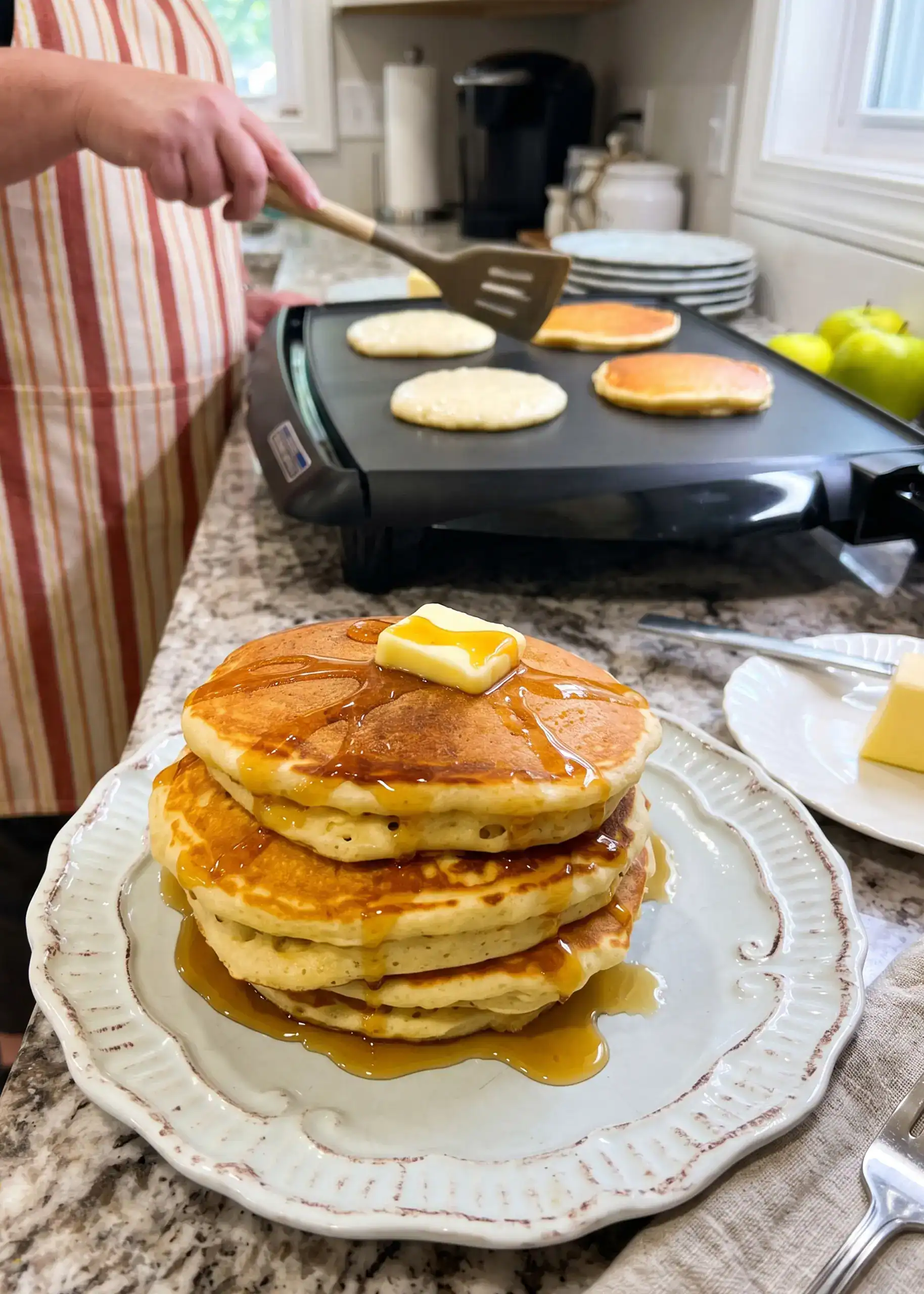 Tall stack of Queen Elizabeth pancakes with butter and maple syrup, with more pancakes cooking on the griddle